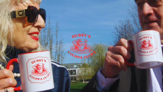 Photo of an older couple drinking tea in the sunshine from mugs that read 'Gussy's Cuppa Club'. (Brent Cross Town)
