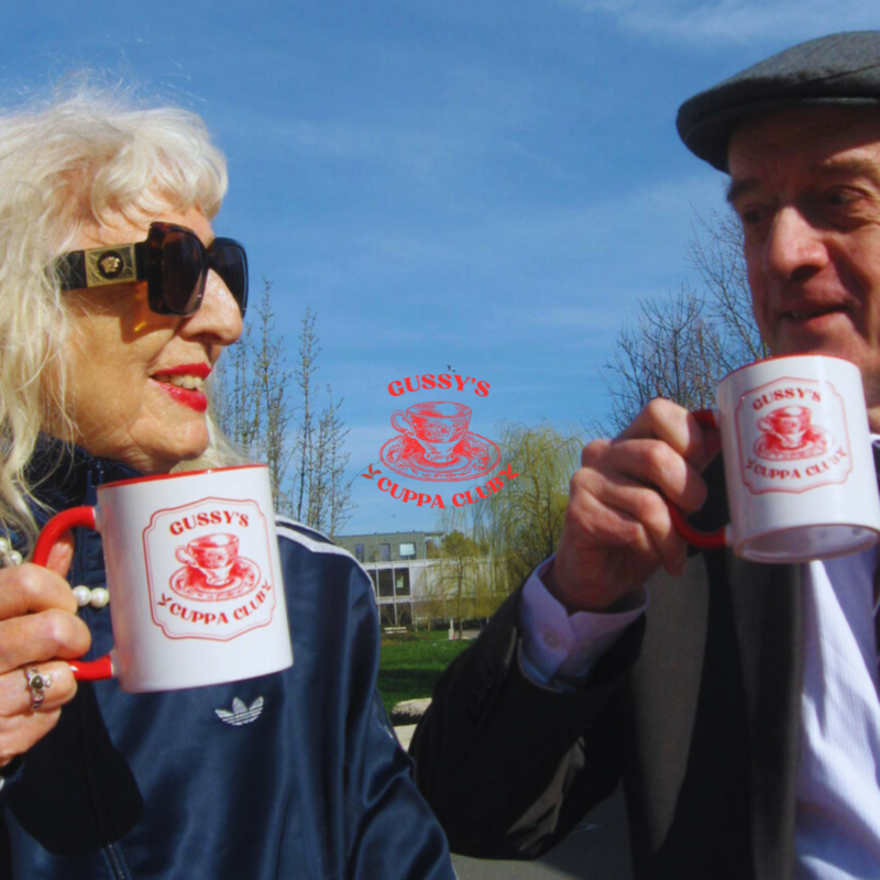 Photo of an older couple drinking tea in the sunshine from mugs that read 'Gussy's Cuppa Club'. (Brent Cross Town)