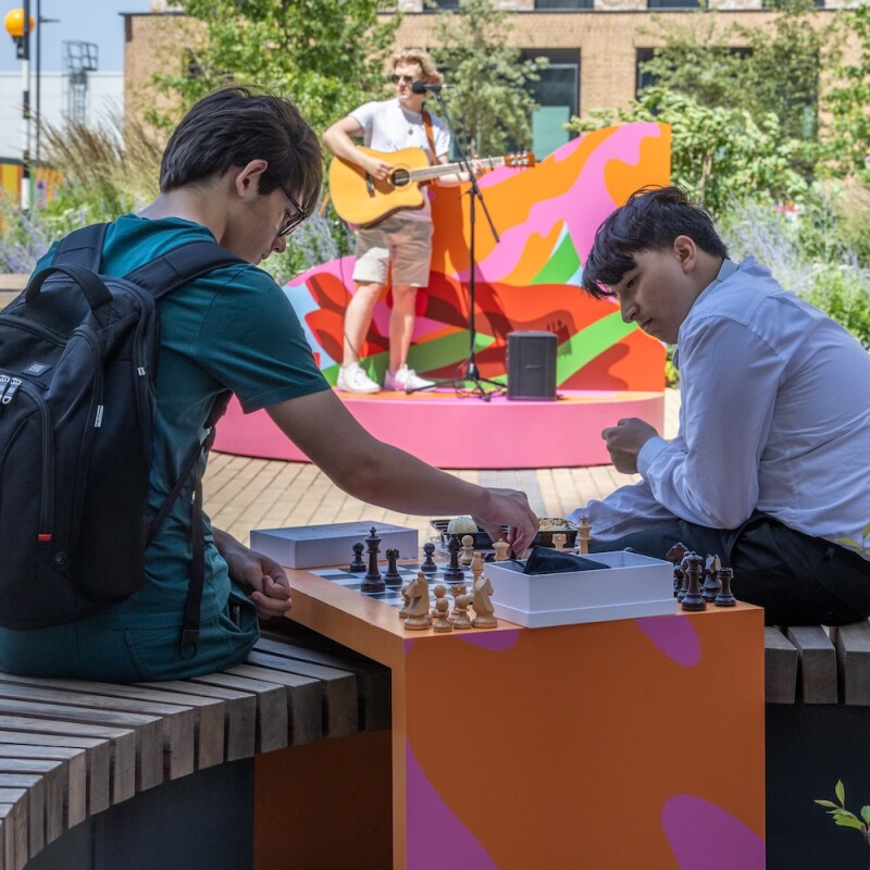 Photo of people playing chess in Neighbourhood Square in Brent Cross Town, while someone performs live music in the background. (Brent Cross Town)